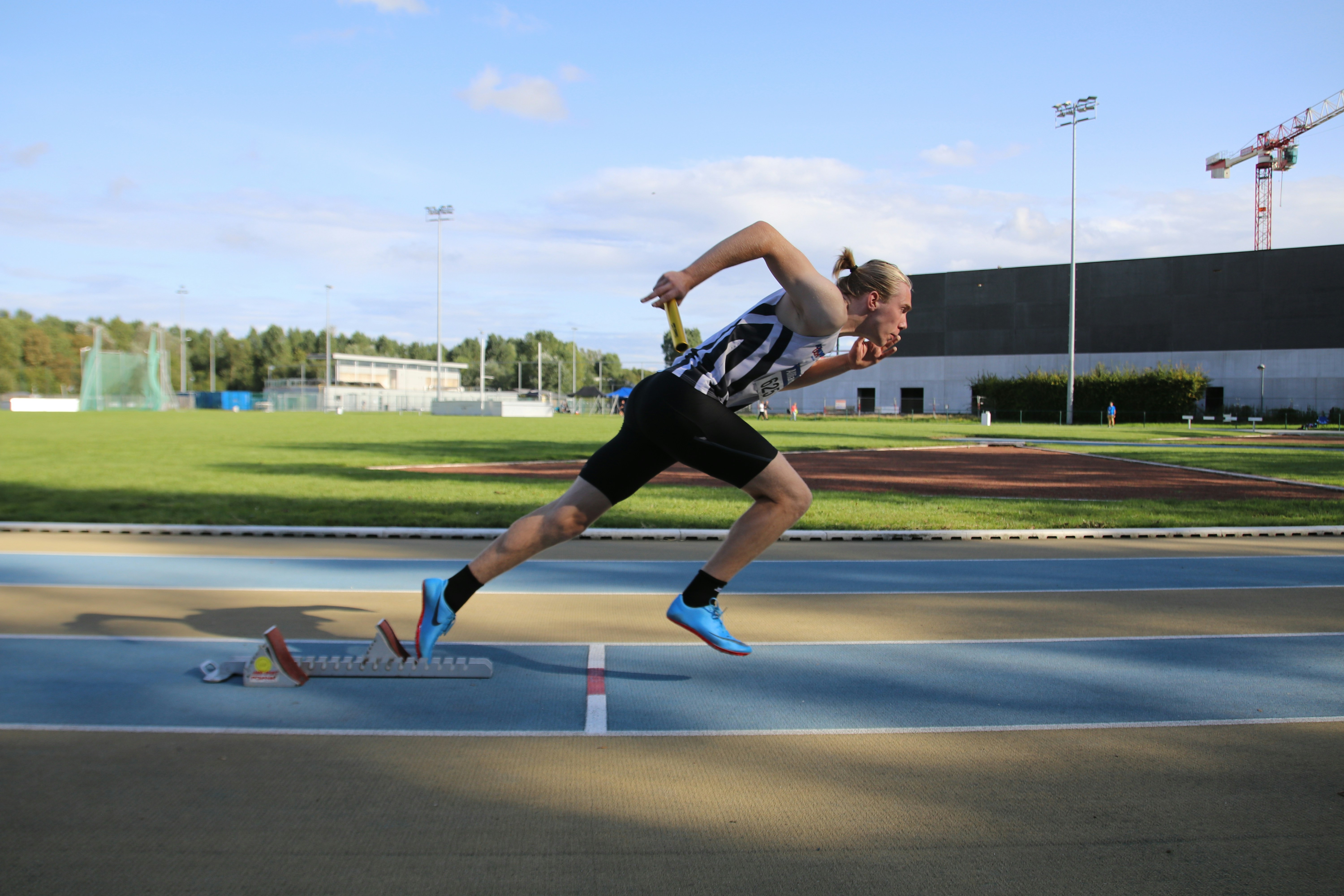 A tall, man with blonde hair in a pigtail and black and white running gear on a running track, pictured launching himself from some starting blocks A tall, blonde man launching from starting blocks