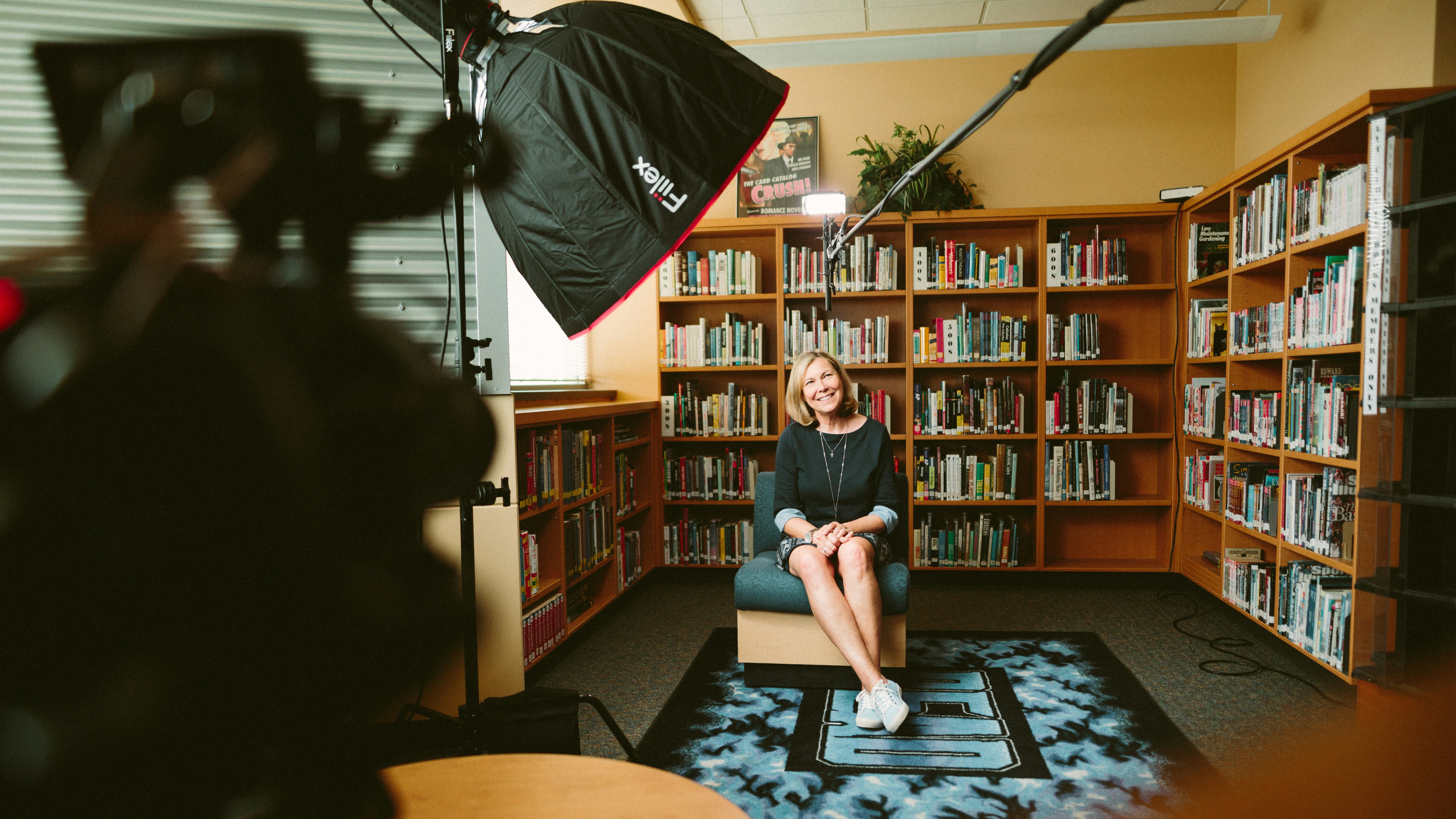 A woman sitting in a chair in center of a room lined with bookcases, an array of camera equipment within radius of her person